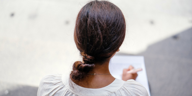 A woman with long, brown hair in a loose bun and light brown skin wears a white shirt with puffy sleeves and a gold necklace. We can only see the back of her body. She leans over a white notebook and writes in it.