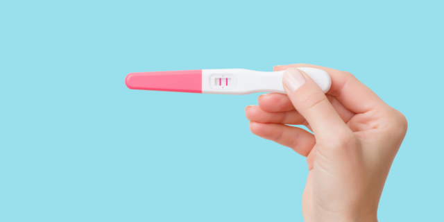 Against a robin's egg blue background, a white person's hand holds up a pink and white pregnancy test, which reveals two pink lines.