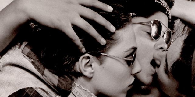 A black and white image shows the faces of three women who are lying on top of each with their arms wrapped around each other. One woman, who wears a bandana around her head and sunglasses, leans in to kiss a woman to the right. On the left, the third woman, who is wearing sunglasses and a denim vest, leans in towards the mouths of the kissing women.