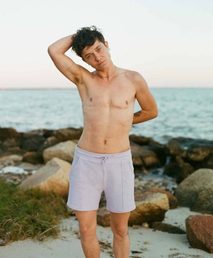 a white trans masc model shows off lavender swim trunks in front of a stony beach