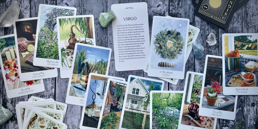 A collection of various tarot cards are strewn on a grey wood tabletop surface, shot from above. In the center is a card with the word "Virgo" in large print and small print description text that is not visible. There are also various crystals.