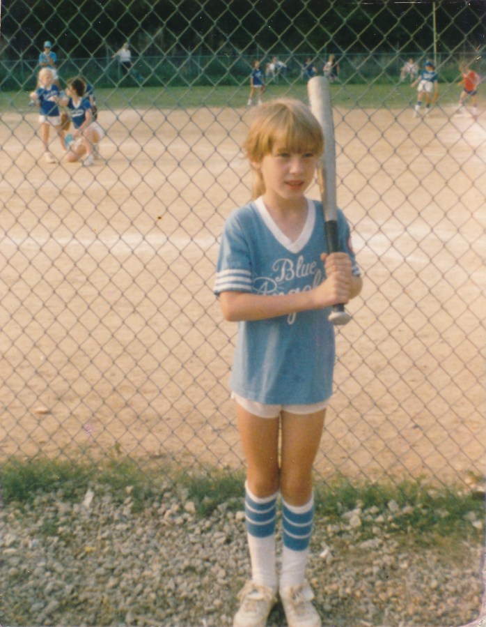 Heather Hogan as a young child holds a bat and wears her Blue Angels softball uniform next to a softball diamond.