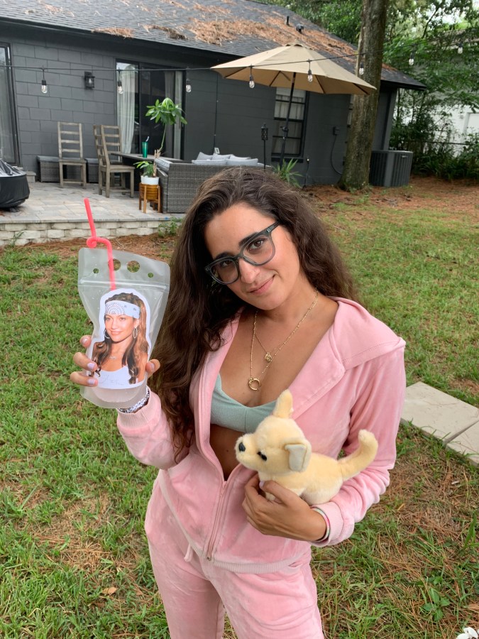 Kayla Kumari Upadhyaya wears a pale pink velour track suit with the hoodie unzipped to reveal a blue sparkly bikini top. She is holding a drink pouch with Jennifer Lopez's face on it in one hand and a stuffed toy chihuahua in the other.