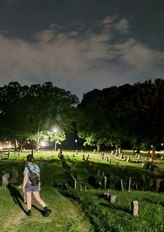 Nico is in a cemetery at night, seen from behind, wearing jorts, black boots and a tropical shirt. They have a backpack and water bottle and shaved dark hair. They are a white genderqueer human.