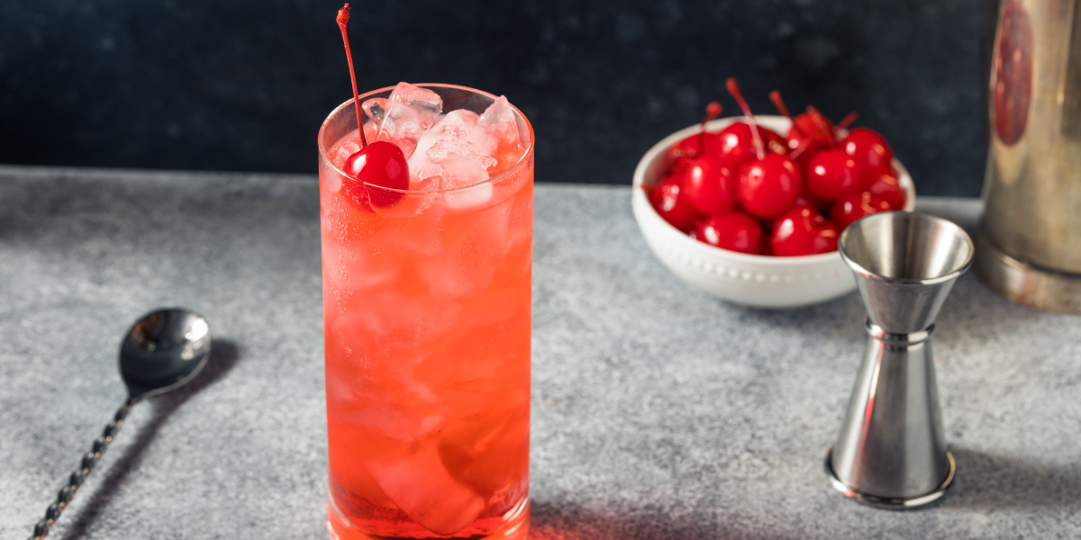 a tall glass of Shirley Temple with ice and a cherry on top. a bowl of cherries is behind it.