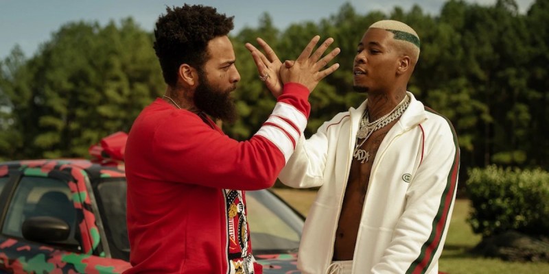 Teak, a light skin black man with a beard, and Murda, a black man with close cut hair bleached blonde, handshake in front of a car