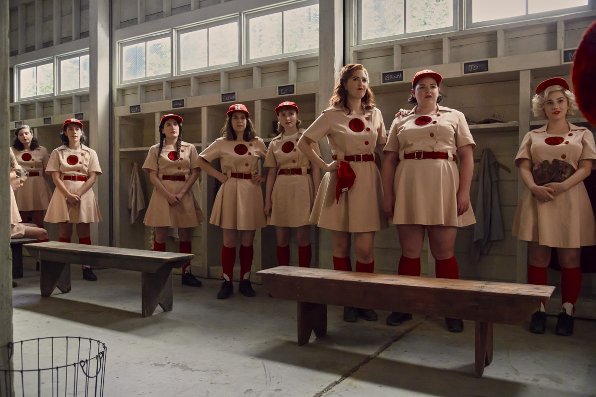 The Rockford Peaches, a team of women from the new series A League of Their Own, stand in a locker room in their skirt uniforms.