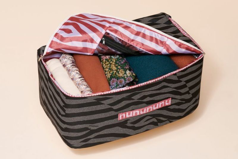 A square duffle bag that is grey and dark brown stripped with a pink stripped inside, it has a pink tag that says "nuuly" across the side, inside the duffle is folded clothes.