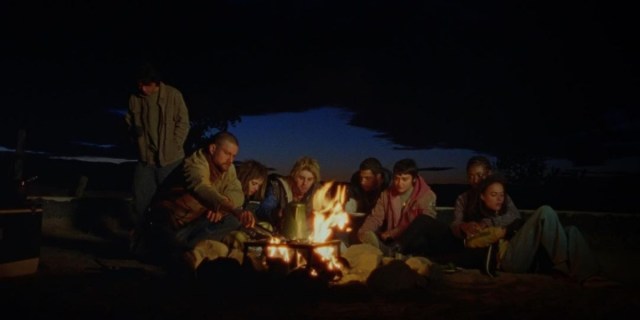 The ensemble of How to Blow Up a Pipeline sits around a campfire. The image is mostly dark. Forrest Goodluck stands behind. Jake Weary, Kristine Froseth, Lukas Gage, Marcus Scribner, and Ariela Barer sit around the fire itself. Sasha Lane leaning on Jayme Lawson sit off to the side.