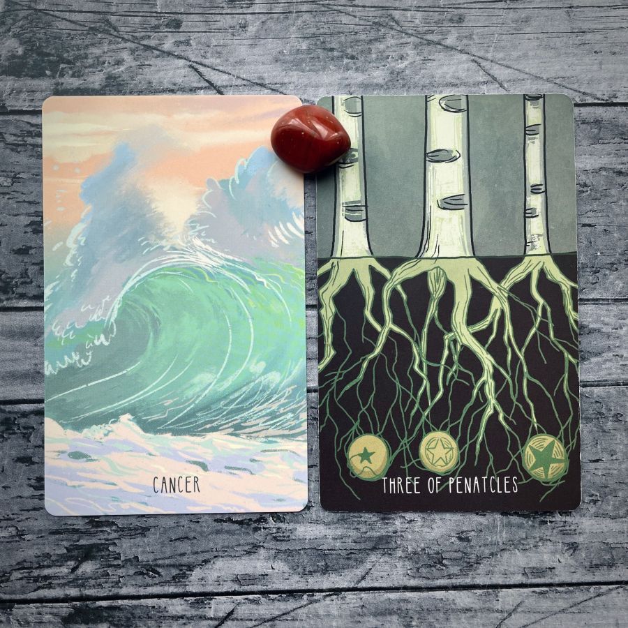 Two cards on a grey wood table:   on the left — a sunset over an ocean wave, it says Cancer at the bottom  on the right — three trees with their roots showing below, it says Three of Pentacles on the bottom