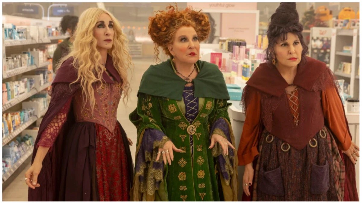 The Sanderson Sisters, left to right: Sarah, who is played by Sarah Jessica Parker, Winifred, who is played by Bette Midler, and Mary, who is played by Kathy Najimi, are standing together in a Walgreens aisle that is lit overhead.