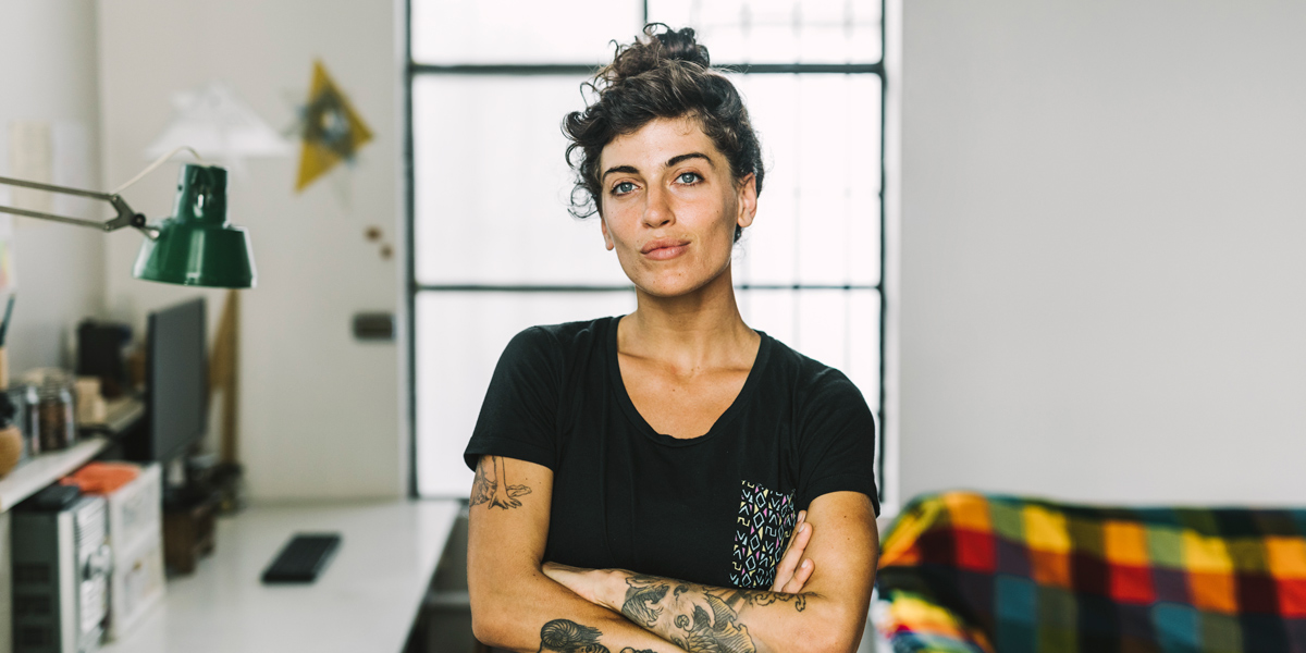 A white queer woman with tattoos has her arms crossed while standing in her workplace, her brunette hair is tied up into a bun.