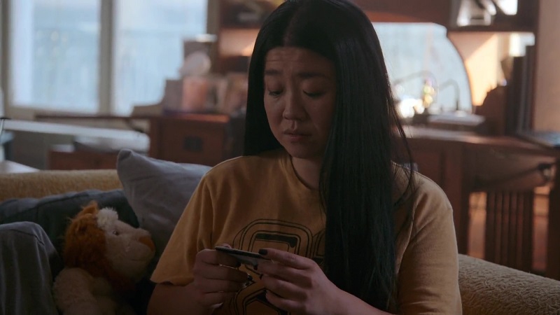 Alice ponders taking Sumi's ID for the audition. In the image, she's sitting on the couch in her loft wearing a mustard colored varsity t-shirt.