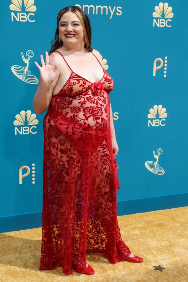 Megan Stalter is wearing a see-through red lace dress with spaghetti straps as she shakes and smiles at the camera