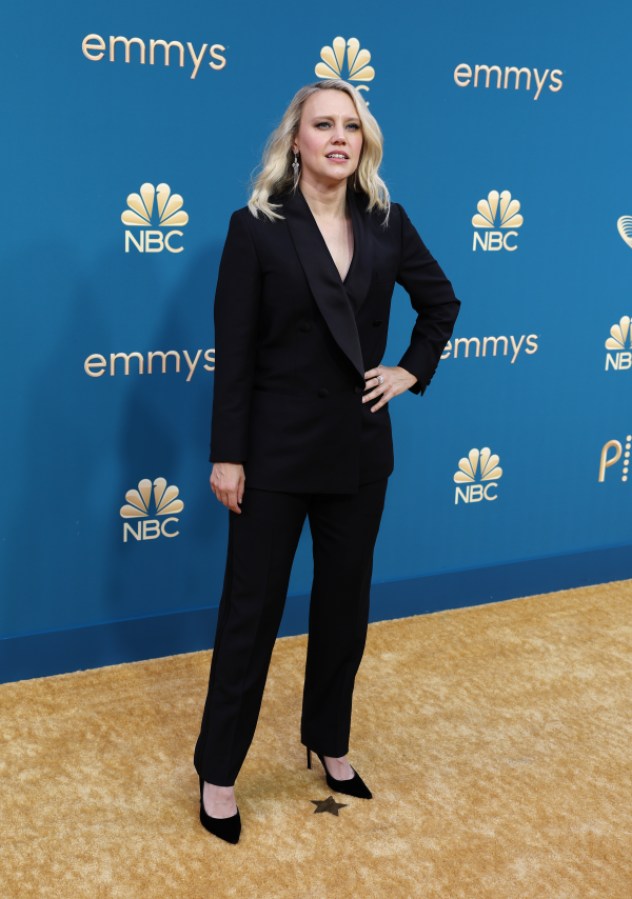 Kate McKinnon is in a black button-up tuxedo with no shirt underneath and simple heels.  She is "smiling" (smiling with eyes, not lips) in front of the camera.