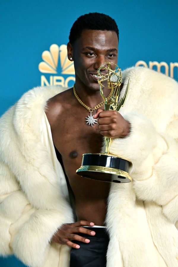 Jerrod Carmichael is shirtless in a long white fur coat holding his Emmy to his face and smiling