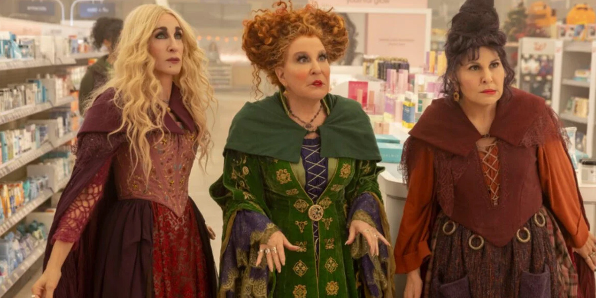 The Sanderson Sisters, left to right: Sarah, who is played by Sarah Jessica Parker, Winifred, who is played by Bette Midler, and Mary, who is played by Kathy Najimi, are standing together in a Walgreens aisle that is lit overhead.