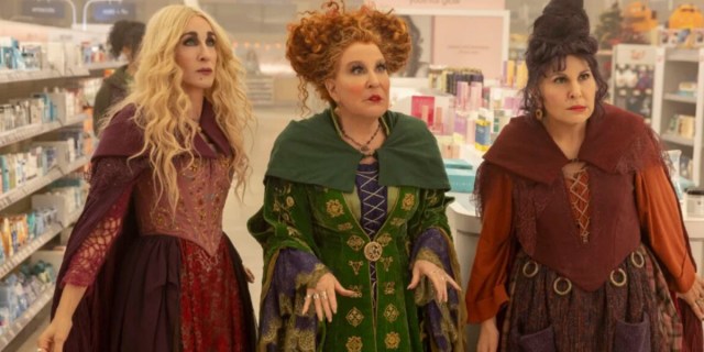 The Sanderson Sisters, left to right: Sarah, who is played by Sarah Jessica Parker, Winifred, who is played by Bette Midler, and Mary, who is played by Kathy Najimi, are standing together in a Walgreens aisle that is lit overhead.