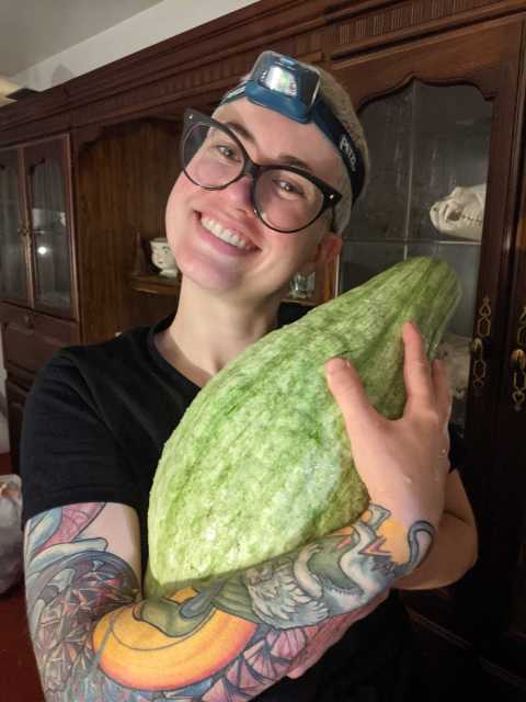 Nico is a white genderqueer human with short, bleached blonde hair and tattoos who is seen here holding a hubbard squash from their garden and smiling broadly. They are wearing glasses and a black tee shirt and a head lamp on their forehead and are in front of a curio cabinet inside their home. It appears to be night.