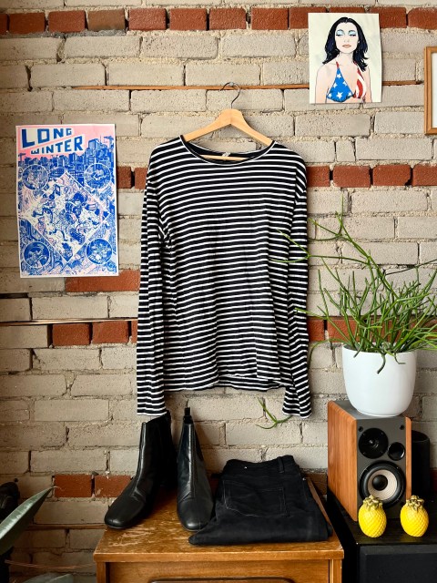 a brick wall, with a photo of PJ Harvey on the upper right corner and a power that says Long Winter to the left. Hanging on a hook is a black and white striped shirt, sitting below is a pair of black jeans and a pair of black boots