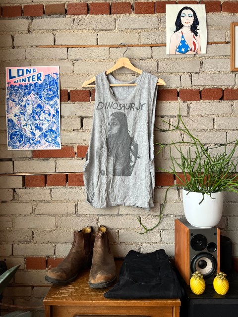 a brick wall, with a picture of PJ Harvey in the upper right corner and a power that says Long Winter in the left.  Hanging from a hook is a gray sleeveless shirt with an image of a girl smoking a cigarette and the text Dinosaur Jr, sitting below is a pair of black jeans and a pair of dirty brown Blundstone boots