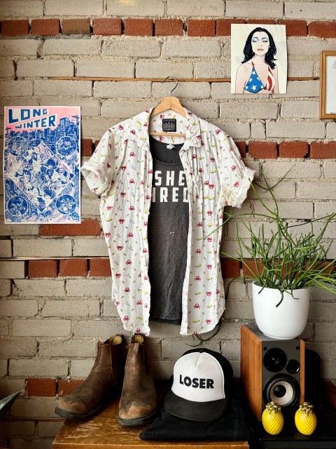 a brick wall, with a picture of PJ Harvey in the upper right corner and a power that says Long Winter in the left.  Hanging from a hook is a gray sleeveless shirt that says She Shreds under a white short sleeve button up with a pattern of lobsters and butter on it, sitting below is a pair of black jeans and a pair of dirty brown Blundstone boots and a hat that says LOSER on it