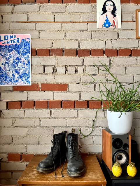 a brick wall, with a photo of PJ Harvey on the upper right corner and a power that says Long Winter to the left. A pair of black Doc Martens sits on a Brown shelf
