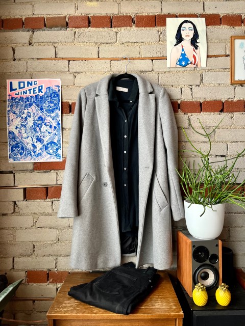 a brick wall, with a photo of PJ Harvey on the upper right corner and a power that says Long Winter to the left. Hanging on the wall is a grey long jacket hanging over a black silk blouse A pair of black jeans sits on a brown shelf