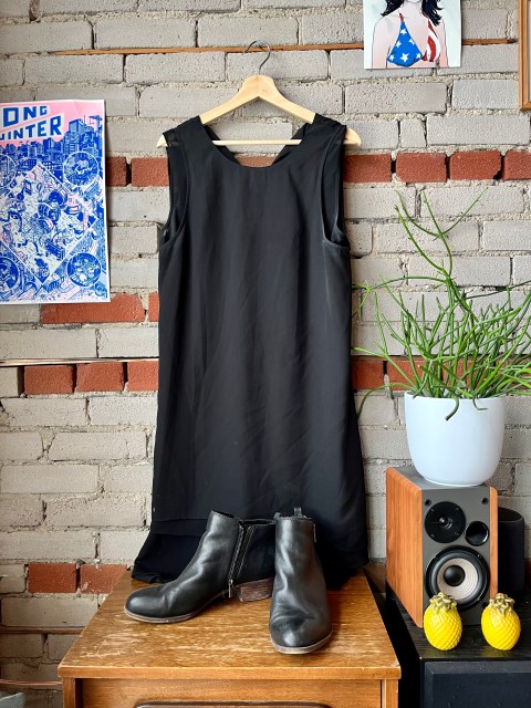 a brick wall, with a photo of PJ Harvey on the upper right corner and a power that says Long Winter to the left. Hanging on the wall is a black sleeveless dress. A pair of black boots sits on a brown shelf