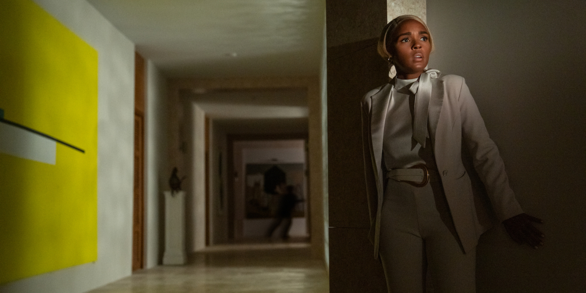 Janelle Monáe as Cassandra Brand hides behind a white wall wearing a white suit. She looks scared.