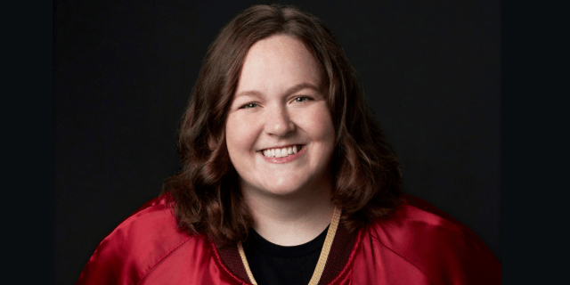 Molly Kearney, Saturday Night Live's first nonbinary cast member, is a white person with shoulder length brown hair wearing a red jacket