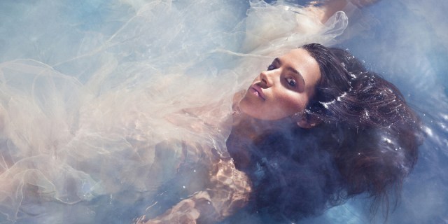 A Latina with long brown hair floats in a wispy pool of water while staring at the camera