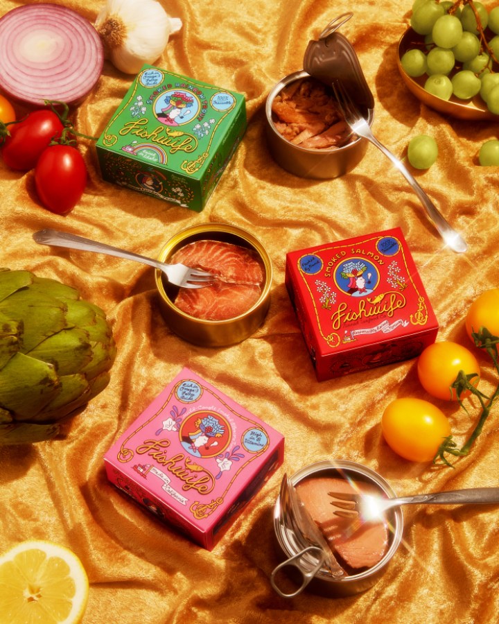a gorgeous display of various fishwife canned fish offerings, tins open, upon a golden velvet background, dotted with fruits and vegetables
