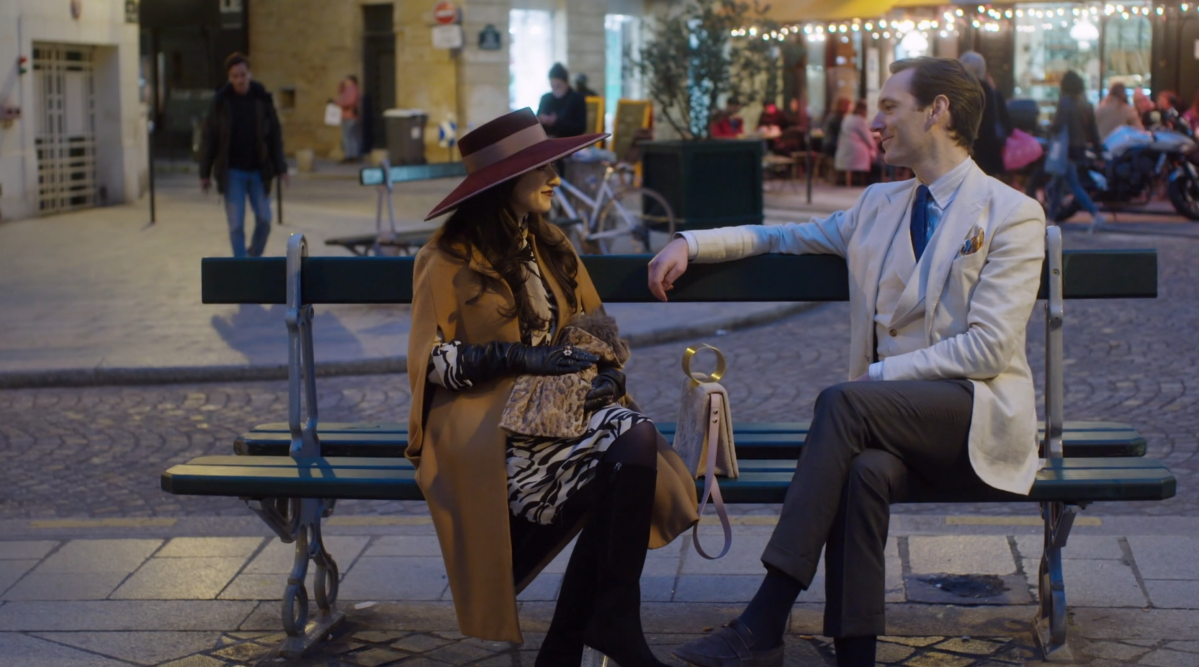 Anya Firestone and her fiancee Matthieu Rasset sit on a bench in the series premiere of Real Girlfriends in Paris on Bravo. Anya is wearing a wide brimmed dark purple hat over a dress with black leggings, black elbow length gloves, and a camel cape coat. Matthieu is wearing a tan three-piece suit.
