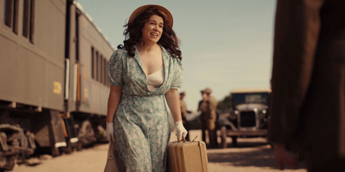 Carson Shaw in A League of Their Own wears a blue dress that's open with her white bra showing and wears white gloves with a sun hat and holds a suitcase while looking frazzled.