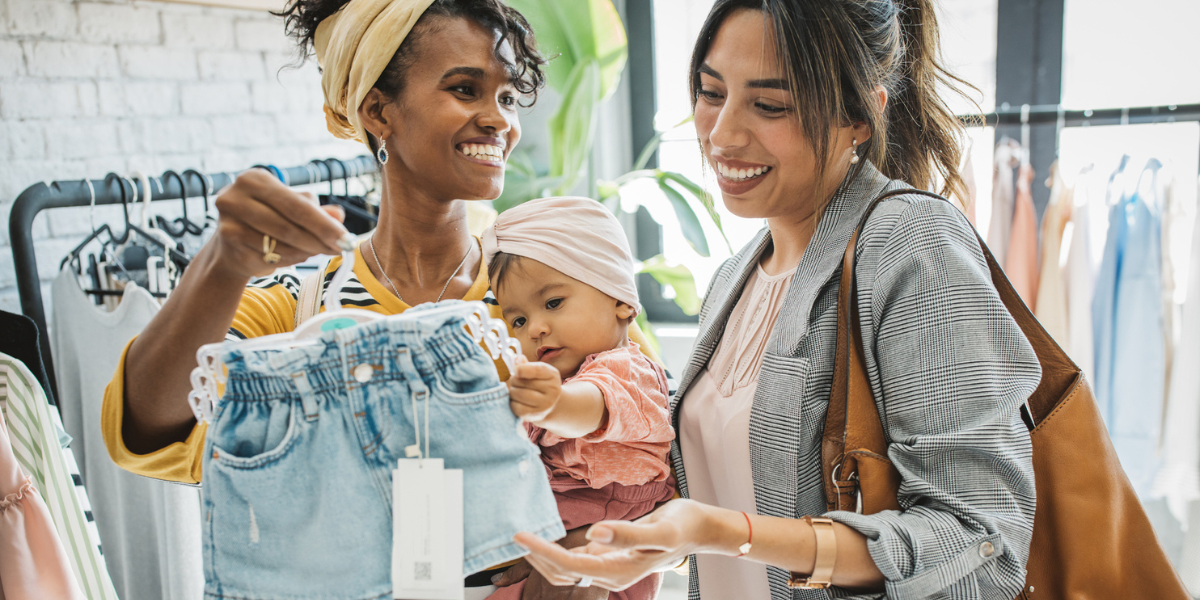 a lesbian couple with an adorable baby shop for clothes in a store. they're smiling at each other and looking at a pair of jorts.