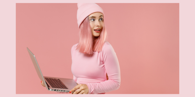 a queer person with pink hair and a pink beanie holds a laptop and looks nervous