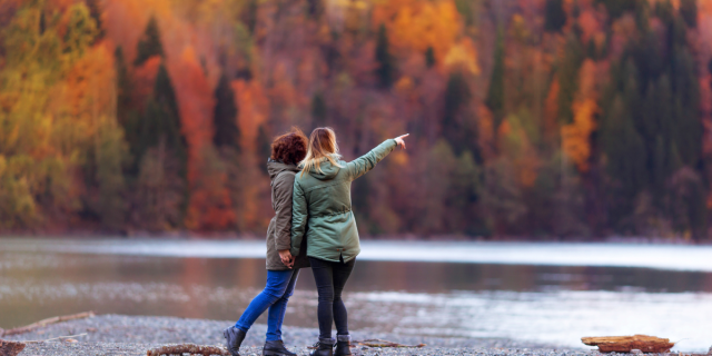 a couple of women hold hands and lean into each other while looking out at a beautiful forest of autumn leaves turning brilliant colors. They are seen from behind and one of them is pointing out at the leaves while the other looks on
