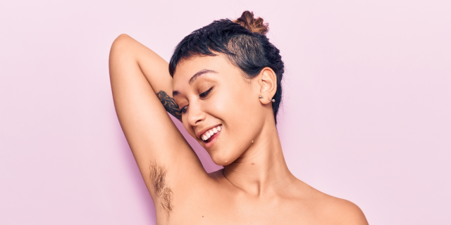 Against a light pink background, a shirtless person with short brown hair and a forearm tattoo holds up their arm, bending at the elbow with their hand behind their head. They look at their armpit hair and smile.