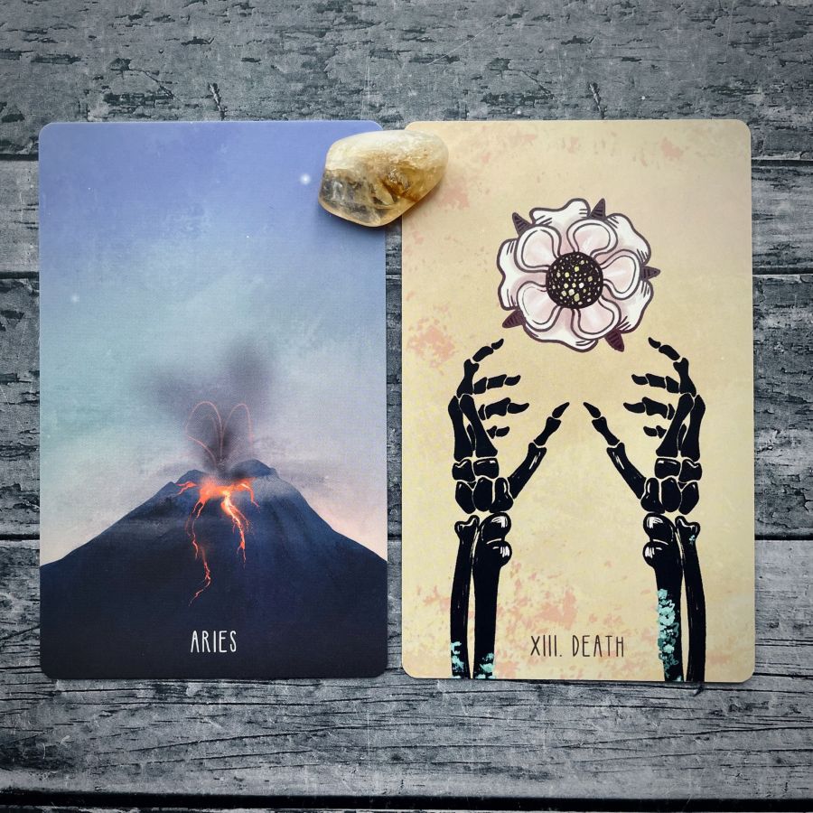 Two cards on a grey wood table:   on the left — a volcano erupting, it says Aries at the bottom  on the right — two skeleton hands and a flower, it says XIII Death on the bottom