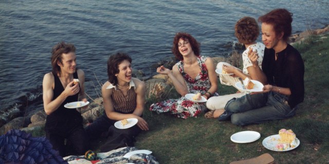 Nan Goldin's Picnic On the Esplanade. A group of five queer friends eat cake from paper plates on a grassy hill. They're all laughing and the ocean is behind them.