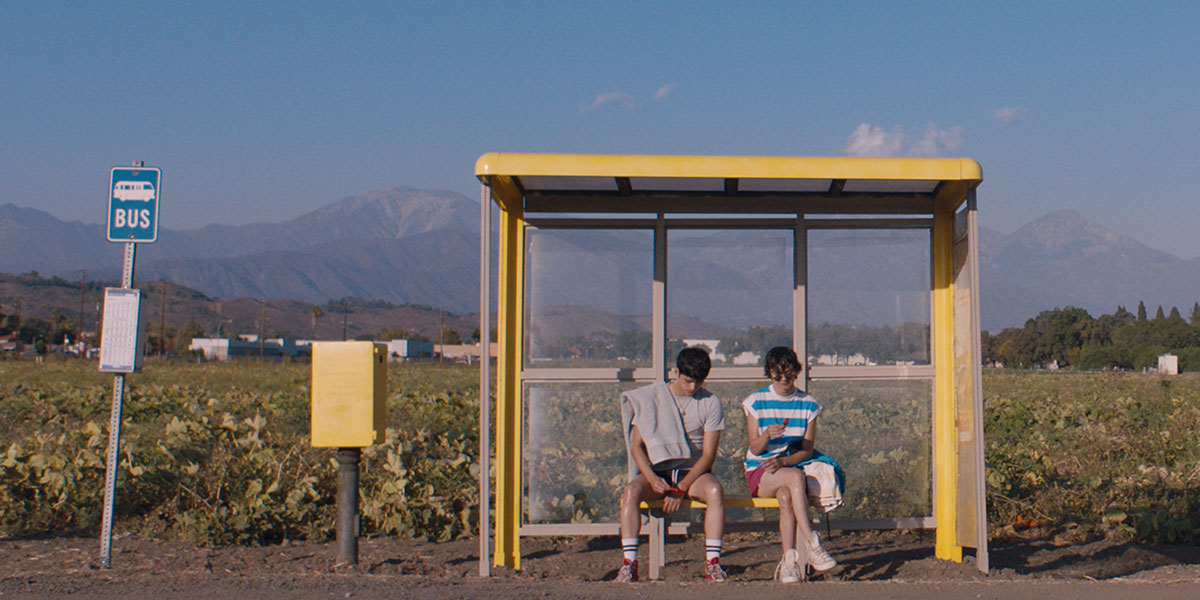 Max Pelayo as Aristotle and Reese Gonzales as Dante sit next to each other at a yellow bus stop with mountains and the sky behind them.