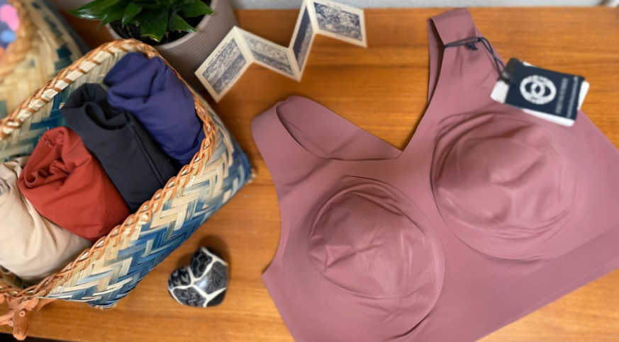 An image of a lavender bra sitting on a wooden table. More bras in various colors are rolled up in a blue and white basket beside it.