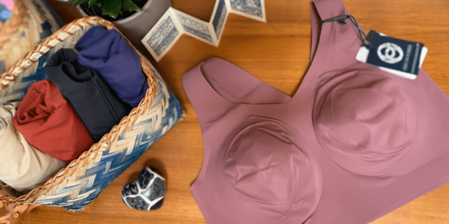 An image of a lavender bra sitting on a wooden table. More bras in various colors are rolled up in a blue and white basket beside it.