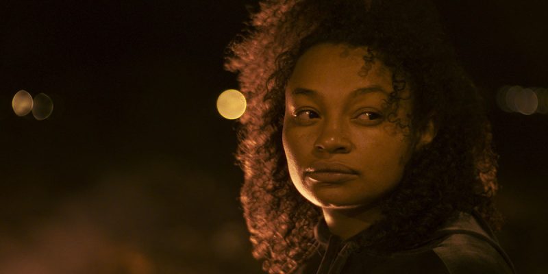 A still from Dry Ground Burning, one of the best 2022 queer festival movies. Close up of Chitarra playing herself. Her face is lit with orange light as she looks off camera.