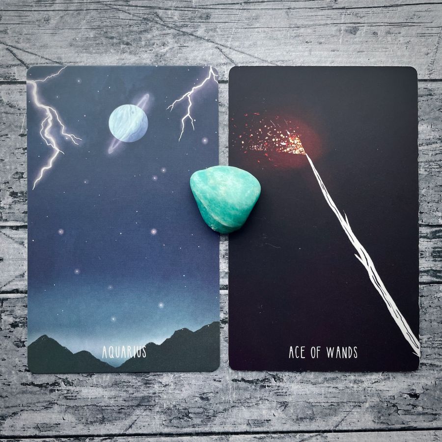  Two cards on a grey wood table:   on the left — a thunderstorm around Neptune at night, it says Aquarius at the bottom  on the right — a fire off of a wood wand at night, it says Ace of Wands on the bottom