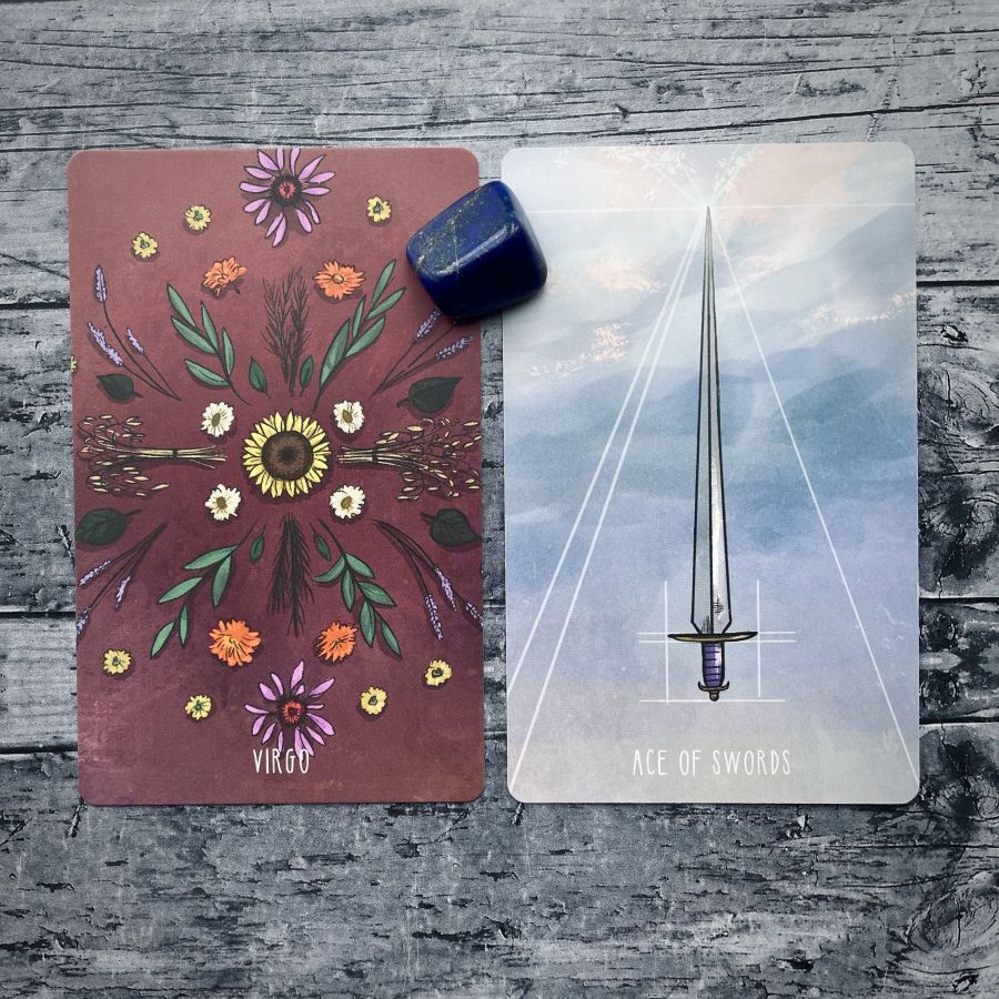 Two cards on a grey wood table:   on the left — various wild flowers make a circular pattern on a rust background, it says Virgo at the bottom  on the right — a sword shining against an abstract watercolor blue background, it says Ace of Swords on the bottom