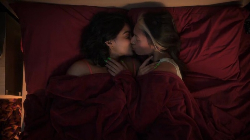 Zaara and Yazmine kiss after hooking up in Zaara's bed. The red covers are pulled up, above their chests. Zaara's wearing a barely visible tank top and Yazmine is in a hot pink bra.
