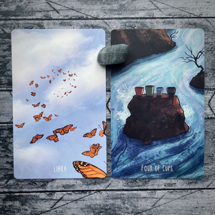  Two cards on a grey wood table:   on the left — a group of monarch butterflies fly through the sky, it says Libra at the bottom  on the right — four cups sit on a rock in a river stream, it says Four of Cups on the bottom