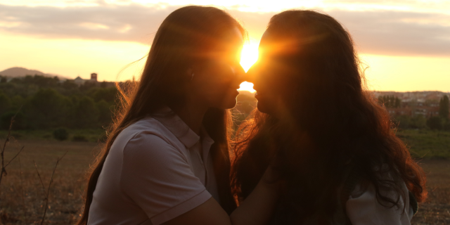 two women with long hair kiss in front of a sunrise that is obscuring their faces with bright golden sunbeams. they are mostly in shadow